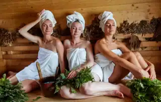 Drei Frauen entspannen in Handtüchern auf einer Holzbank im Wellnessbereich.