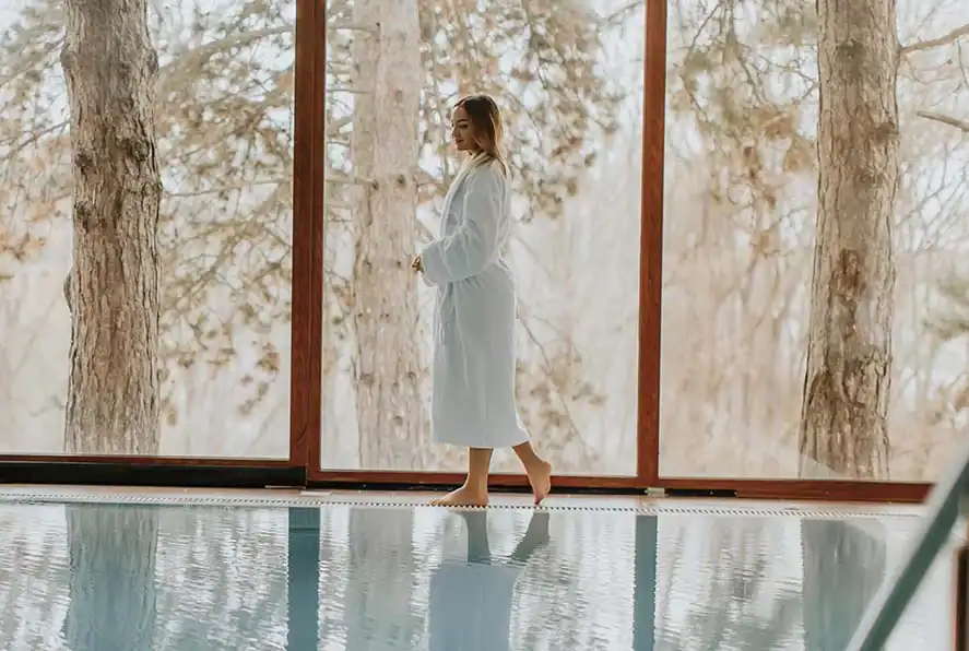 Frau im Bademantel am Pool mit Blick in die Natur.